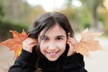 retarto de niña morena sonriente mirando a cámara y jugando con hojas de otoño  en un parque