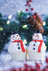 Two iced Christmas Snowman cupcakes with carrot nose, Santa hat, and scarf. Selective focus with blurred foreground and background.