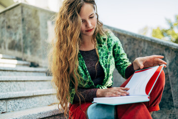 Pretty student female reading a book while sitting on the stairs of the college campus on a sunny...