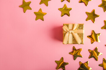 Fototapeta na wymiar Gold box with a gift on a pink background. Gold stars confetti on a pink background. Golden gift and confetti close-up and copy space.