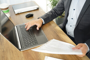 a male businessman is typing on a laptop, looking at the text in a paper document
