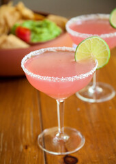 Pink Prickly Pear Margarita with Salt Rimmed Stemmed Glass with Lime Wedge on Wood Table with Chips, Salsa and Guacamole