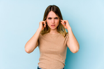 Young caucasian woman isolated on blue background covering ears with fingers, stressed and desperate by a loudly ambient.