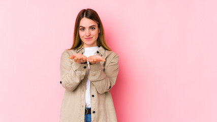 Young caucasian woman isolated on pink background holding something with palms, offering to camera.