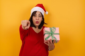 Indoor shot of cheerful middle aged woman wearing Santa hat  indicates happily at you, chooses to compete, has positive expression. beautiful clever student makes choice, isolated on yellow Wall