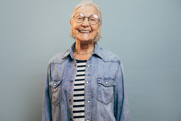 Cheerful very emotional old woman with gray hair wearing jeans jacket and shirt looking at camera, laughing and posing isolated over blue background. Smart happy senior lady. Happy feelings concept.