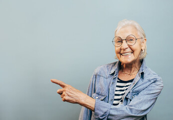 Nice friendly old woman with white hair and round glasses wants to show something to you. Stylish old ages concept. Isolated over blue background. Jeans jacket and striped shirt.