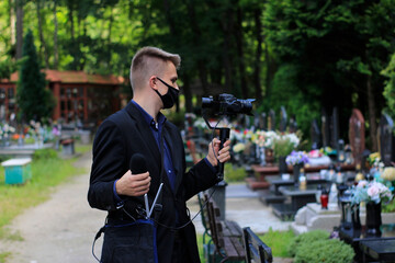 Elegant man in suit is recording grave with cross full of flowers. Funeral live streaming on cementary. Cameraman handing a microphone and camera on a gimbal.
