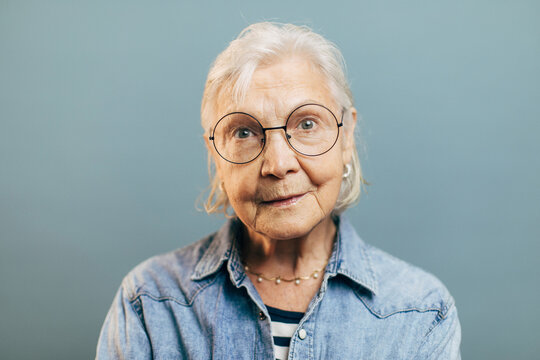 Wise concentrated old woman with attentive gray eyes in big round glasses looks into camera. Intelligent elderly lady wearing striped shirt and denim jacket isolated over gradient blue background.