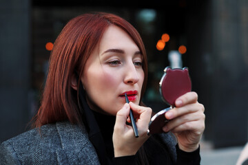 Pretty redhead woman looks at pocket mirror and applys red lipstic before business meeting. Dating after work concept.