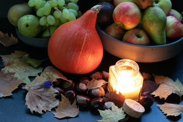 Organic orange pumpkin in a composition with grapes, peers, apples, autumn dried leaves, wine, candle light in a dark marble background. Harvest and cold times
