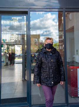 An Elderly Woman In A Black Jacket And A Black Medical Mask Walks Out Of The Door Of A Shopping Center.