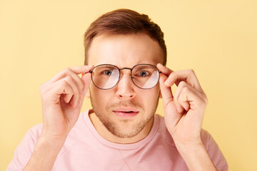 Portrait of caucasian man keeps hands on rim of spectacles, funny reaction to shocking news, gaze at the camera isolated on yellow background in casual clothes