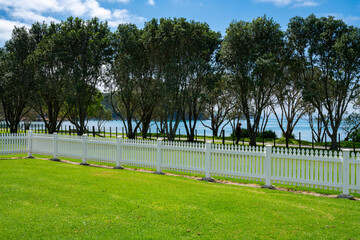 white picket fence across green lawn with row of pohutukawa trees and Man O' War Bay behind.