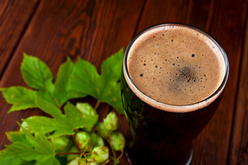 Glass of dark stout beer with natural green hop leaves on a wooden table. 
