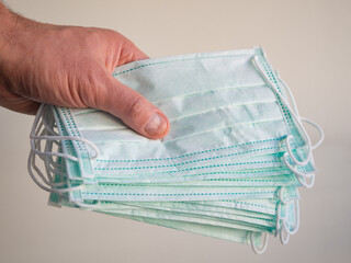 A lot of medical or surgical green color masks in the hand of a young man, isolated on a light-colored wall background. The concept of medical supplies or accessories for personal protection. Close-up