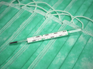 Close-up of many stacked medical or surgical green color protective masks with ear loops as background. Glass mercury thermometer on a masks. Concept of supplies or accessories for personal protection