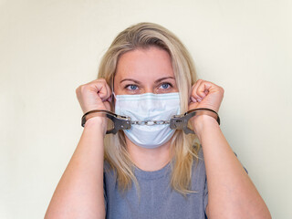 Young girl in a mask with handcuffed hands . Handcuffs are put on raised hands. The model is isolated on a light-colored wall background. Quarantine and freedom restriction concept. Close-up