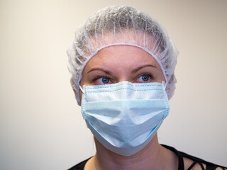 Young girl in a medical surgical blue color mask. On the head is a protective cap or hat. The model is isolated on a light-colored wall background. Quarantine and freedom restriction concept. Close-up