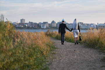 back view of newlyweds walking away to the river. Autumnal landscape and cityscape on background. Modern Groom and bride. Birds flying. Sunny evening. Blue sky. Fibonacci spiral image view.