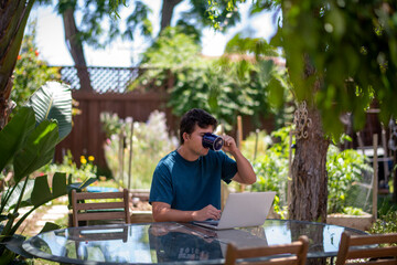 young adult male drinking coffee, working on computer outside backyard