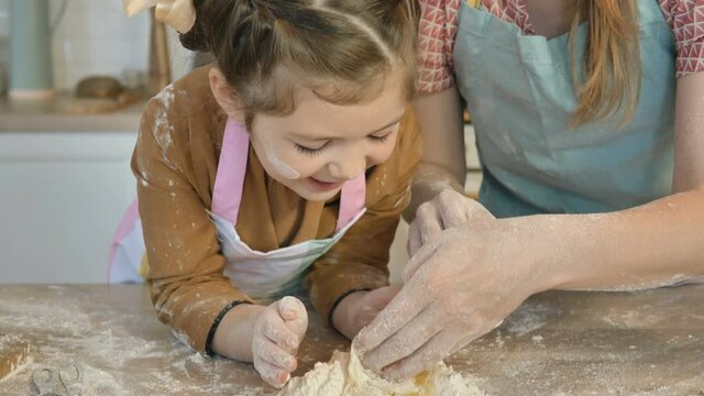 Smiling mother helps little daughter to mix egg with flour making pie dough at large wooden table in light kitchen closeup