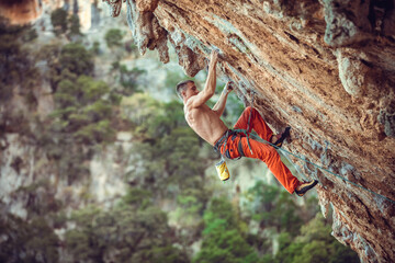 Young male rock climber on overhanging wall. Rock climbing on natural cliff. Strong young man struggling up challenging route.