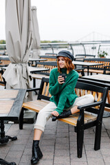 Portrait of a beautiful red-haired girl.  Beautiful red-haired girl in a green sweater and cap sits with a cup of coffee in a cafe on the street.