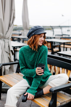 Portrait of a beautiful red-haired girl.  Beautiful red-haired girl in a green sweater and cap sits with a cup of coffee in a cafe on the street.
