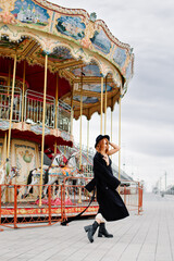 A beautiful red-haired girl in a black coat and hat stays near the merry-go-round.