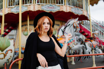 Fototapeta na wymiar A beautiful red-haired girl in a black coat and hat stays near the merry-go-round.