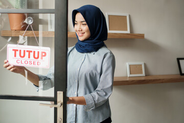 cheerful woman in closed sign and smiling while standing against front door office studio. Tutup in english mean close