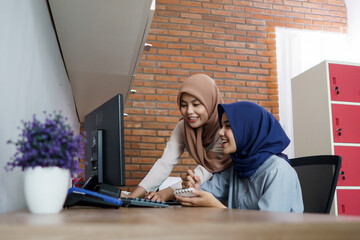 attractive cheerful young muslim business woman working on computer and smiling while sitting at her desk modern office with her friend at studio