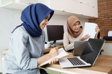 attractive cheerful young muslim business woman working on laptop and smiling while sitting at her desk modern office with her friend at studio