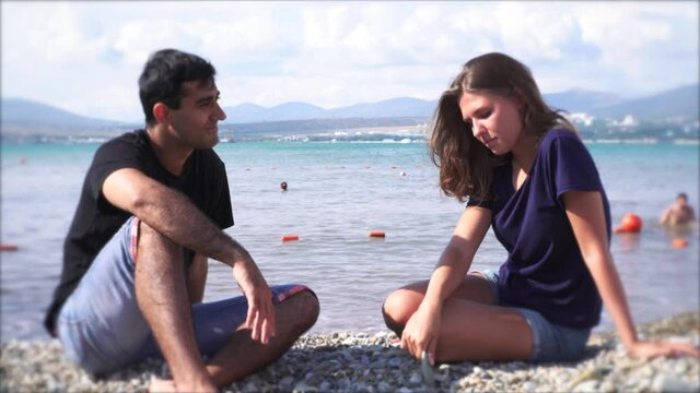 Young couple having an argument by the sea. Media. Man and woman sitting on a stony sea shore, woman feeling ashamed, looking down and touching stones.