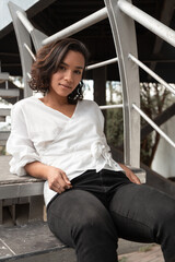 Latin young brunette woman with curls and short hair sitting relaxed on stairs, wearing a white blouse and black pants, photo in the day