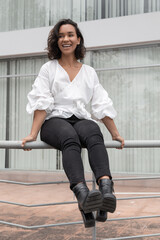 beautiful young latin brunette woman with curls and short hair having fun, sitting on a handrail, wearing a white blouse and black pants, photo in the sunny day outside a building