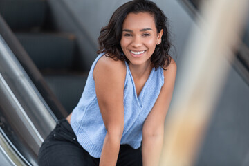 beautiful young brunette latina woman with short hair and curls, smiling sitting on a handrail of electric bleachers, wearing blue blouse and black pants