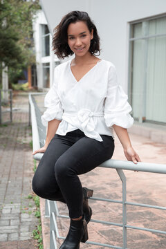 Beautiful young Latin brunette woman with curls and short hair close up, sitting on a handrail, wearing a white blouse and black pants, photo in the day with sun outside a building