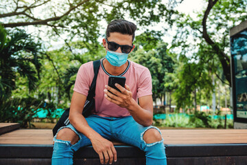 Portrait of a handsome urban man sitting outdoors, using his mobile phone, wearing a mask