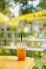 Close-up of cool peach tea in clear plastic glass on a wooden table on a sunny day. The blurred background of yellow, white, and green color with bokeh in a soft tone. There is a copy space on top.