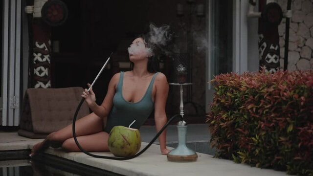 Young European woman with black hair in swimsuit smokes hookah or shisha. Evening rest in an outdoor cafe near swimming pool. Girl inhales and exhales smoke.