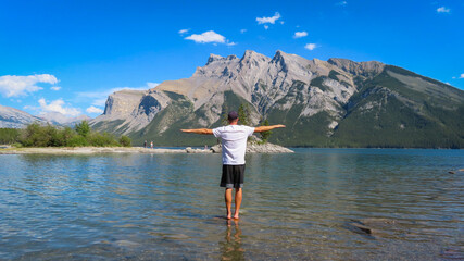 Man standing on the edge of rocky cliff spreading arms towards the beautiful waters of Minnewanka lake in Rocky mountains -  Banff National Park, AB.