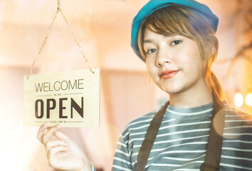 Business owner attractive young Asian woman in apron hanging we're open sign on front door smiling welcoming clients to new cafe. open shop after covid 19 crisis .