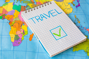 Check mark in box` "Travel" in notepad against colorful world map background. Post-pandemic travel concept. Allowed to travel