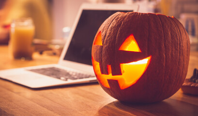 Lonely evening on Halloween. Laptop and lit pumpkin candle at home on the table. Concept of...