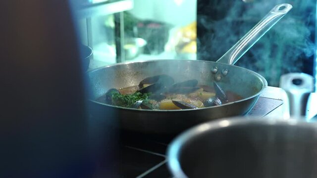 Restaurant kitchen, cafe eats food. Chef cooks sea food, mussels in frying pan. Adds chopped greens from tray, container. Strong steam, smoke, working atmosphere cooking, teamwork. Social media videos