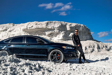 Handsome man in fashion leather jacket and sunglasses stands near black car on white rocks background. Luxury black car.