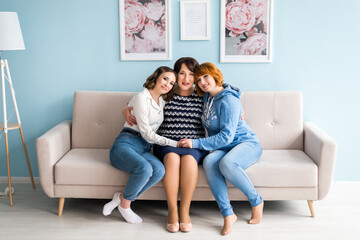 Granny, her daughter and granddaughter are looking at camera and smiling while sitting on sofa at...
