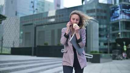 A young woman is talking on the phone while walking and eating a sandwich. Junk food as part of a busy office worker's schedule